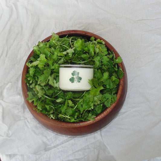 Candle with a cilantro design in a wooden bowl filled with green leaves on a white fabric background