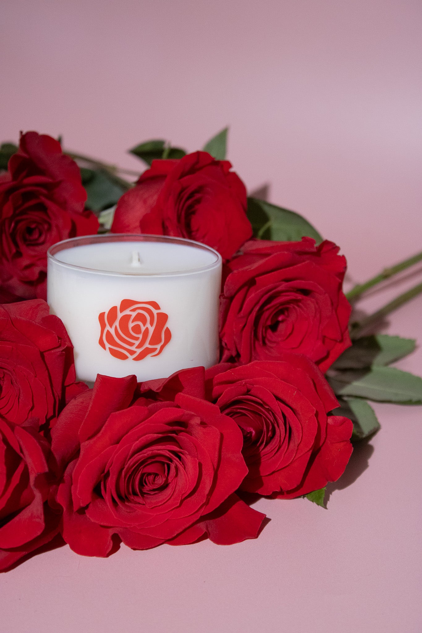 White candle with a red rose logo surrounded by red roses on a pink background