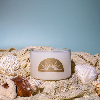 Candle with a sun design surrounded by seashells on a textured surface with a blue background