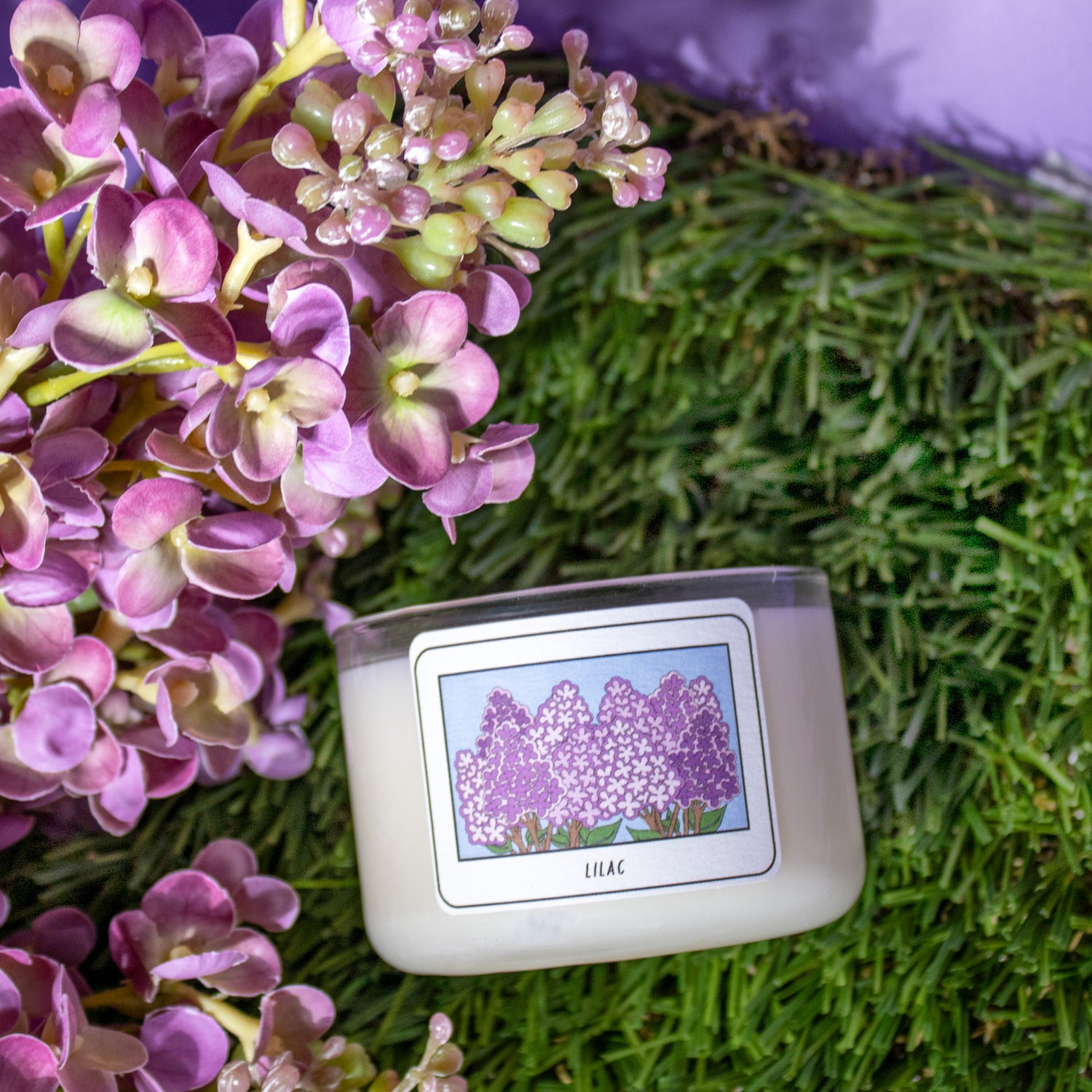 Candle with a lilac design on its label surrounded by purple flowers and greenery