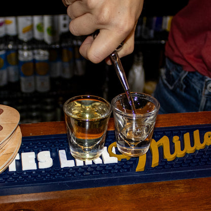 Two shot glasses being filled with a clear liquid on a bar with 'Miller' branding.