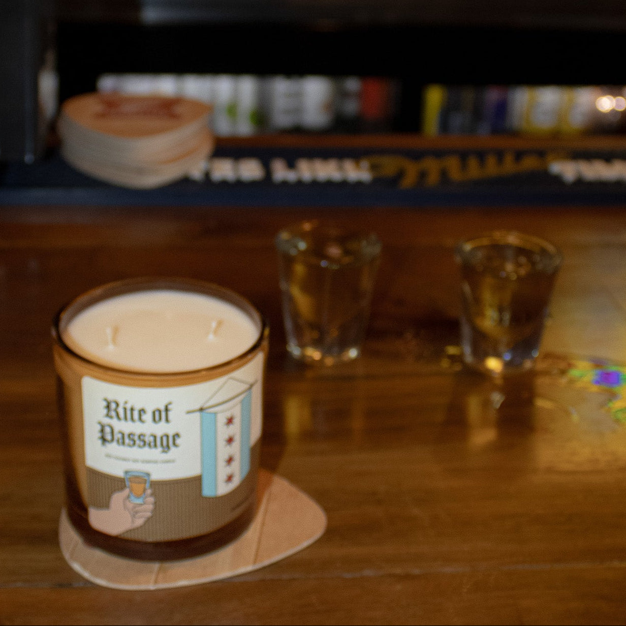 Candle labeled 'Rite of Passage' on a coaster with a bar setting in the background.