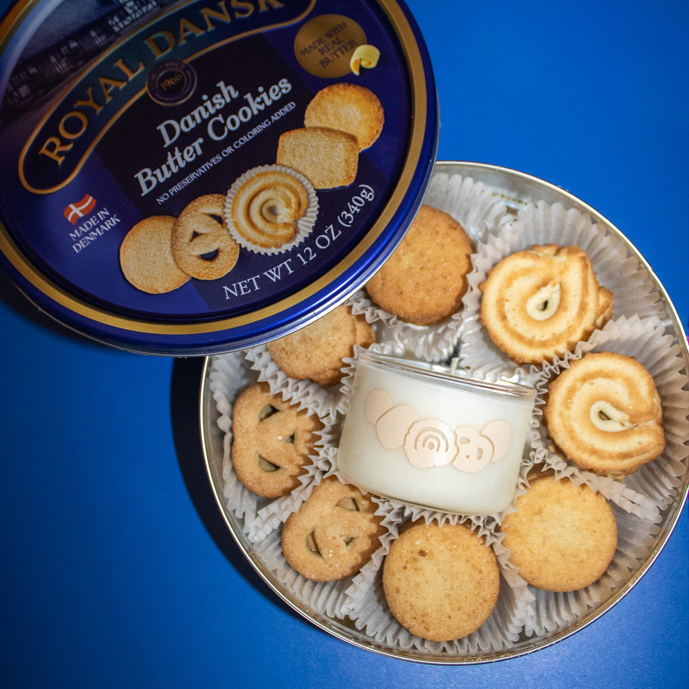 An open round metal tin of Royal Dansk Danish Butter Cookies on a bright blue background, with the lid leaning beside it. The cookies are arranged inside a clear plastic tray, including swirls, striped patterns, and solid round varieties, with a small white wax seal in the center.