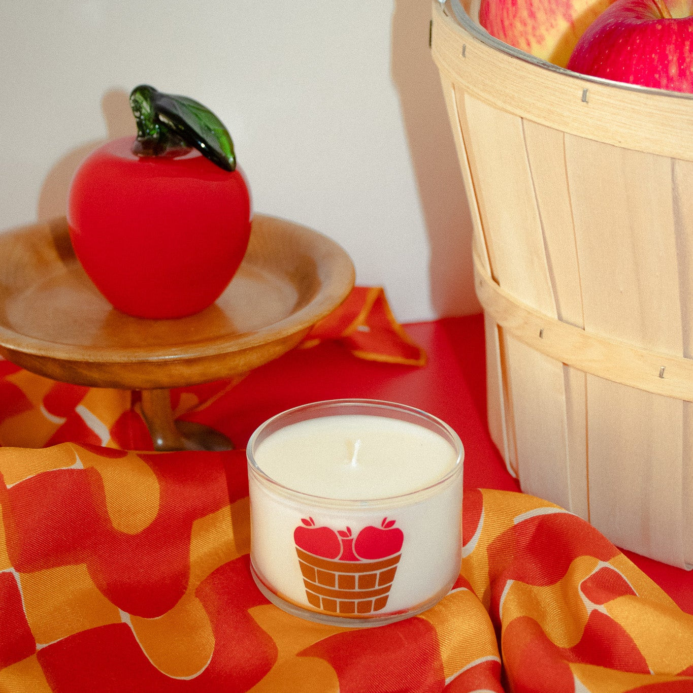 A white candle in a clear glass jar with a red apple and basket graphic, styled on a red and orange patterned fabric next to a red apple sculpture and a wooden basket of red apples.
