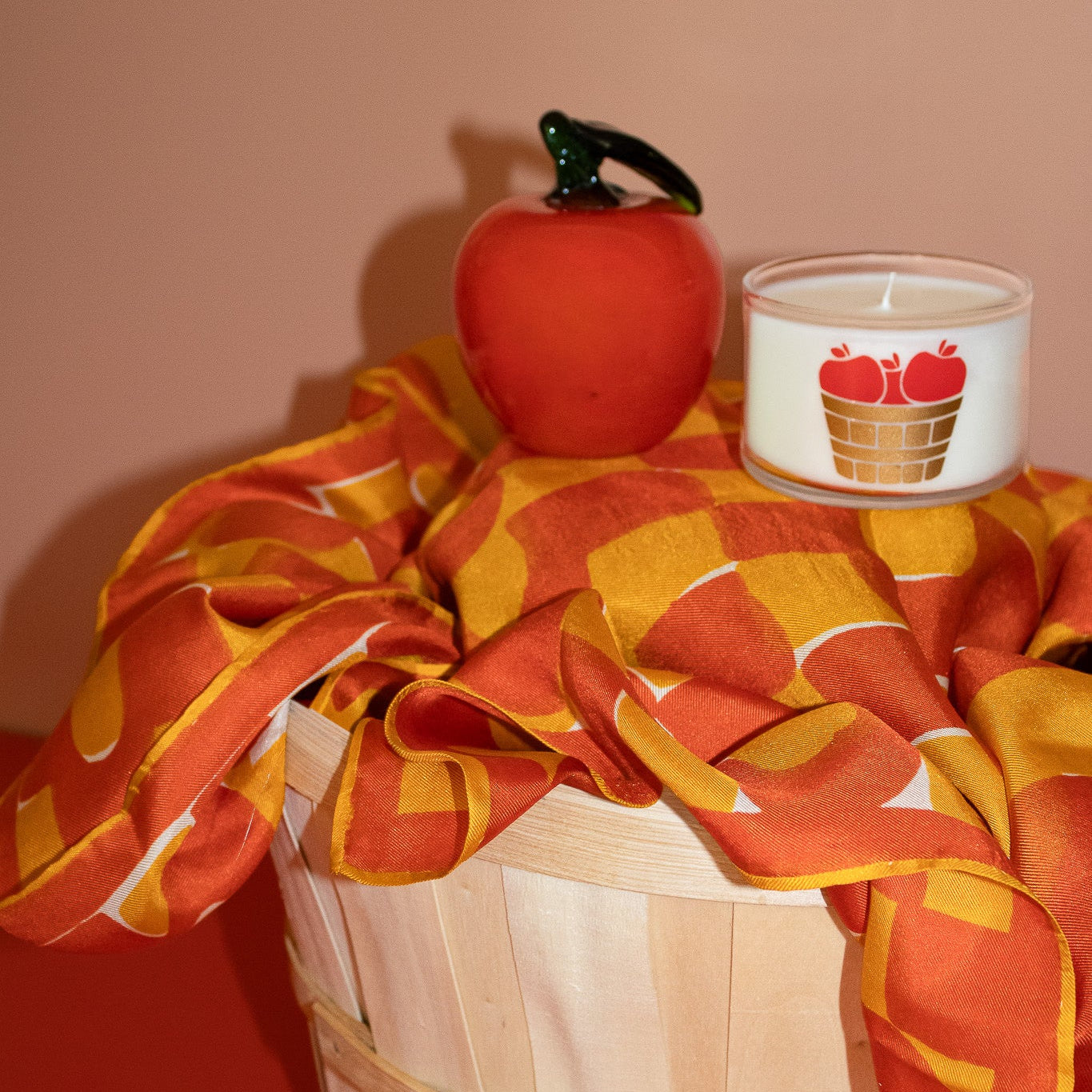 A white candle in a clear glass jar with a red apple and basket graphic on the label, styled with a red apple prop and an orange and yellow patterned fabric draped over a wooden basket against a solid peach background.
