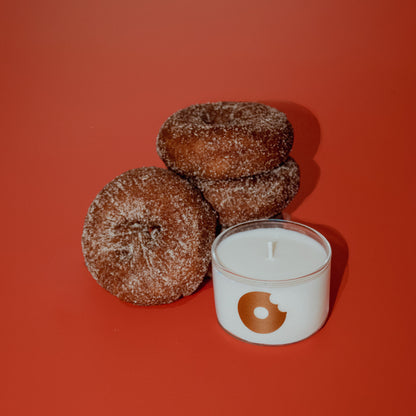 Three sugar-coated donuts next to a white candle in a clear glass jar with a brown donut-shaped logo, set against a solid red-orange background.