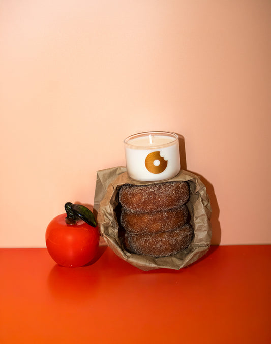 A white candle with a gold donut logo sits on top of a paper bag holding three chocolate-covered donuts, styled next to a red tomato on a warm orange-red surface against a peach background.