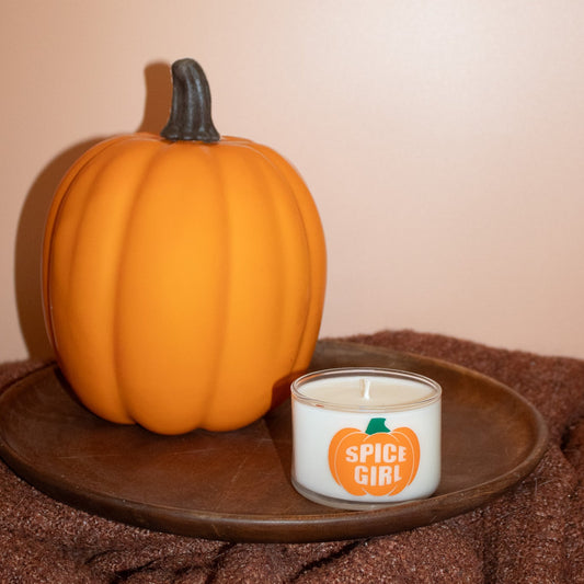 A small white candle in a clear glass jar with a pumpkin graphic and the words 'SPICE GIRL' sits on a round dark wooden tray next to a large orange pumpkin decoration, set against a warm beige wall and brown textured fabric.