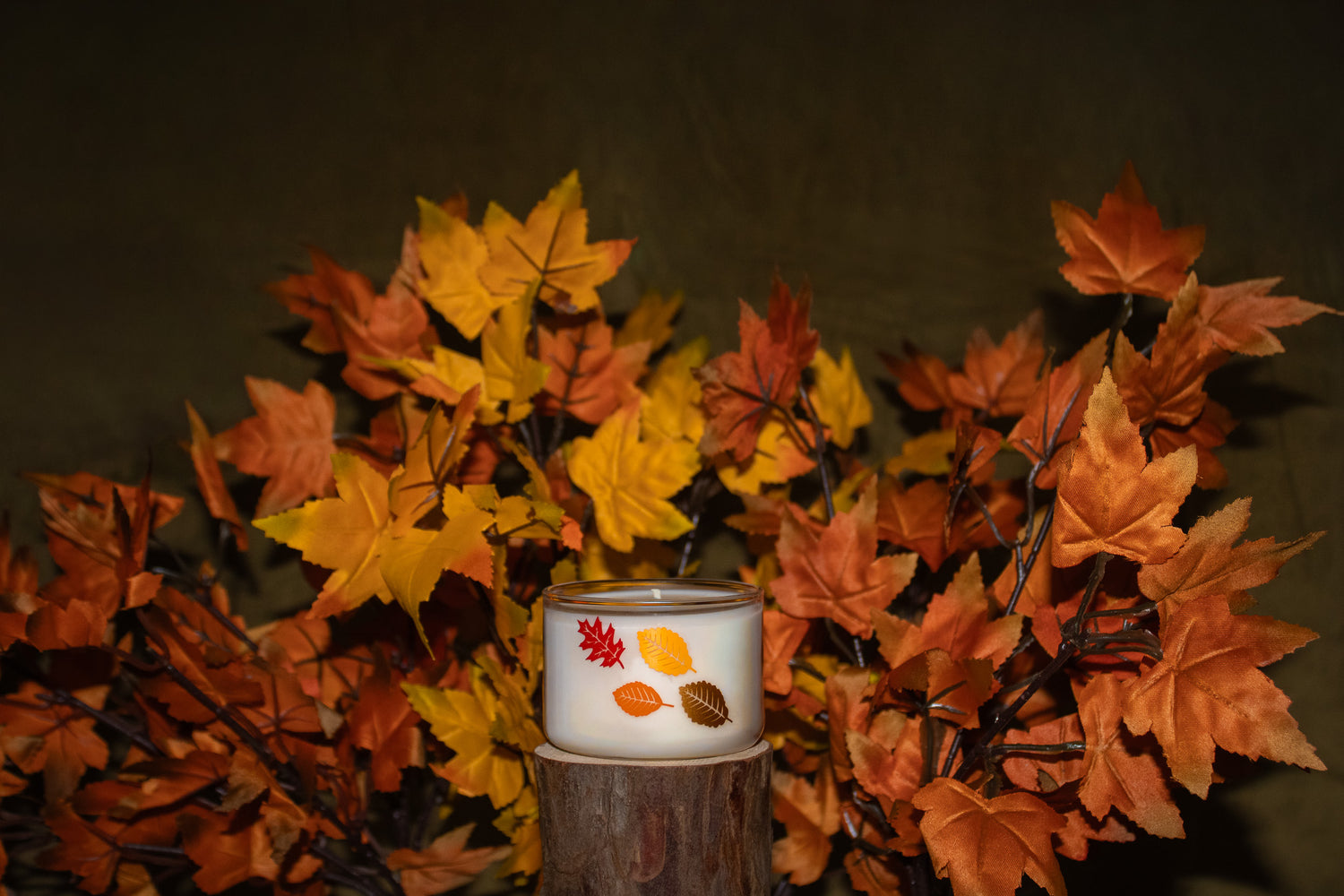 A white jar candle with a gold rim and a printed design of red, yellow, and brown autumn leaves, displayed on a wooden stump prop surrounded by artificial orange and yellow maple leaves against a dark background.