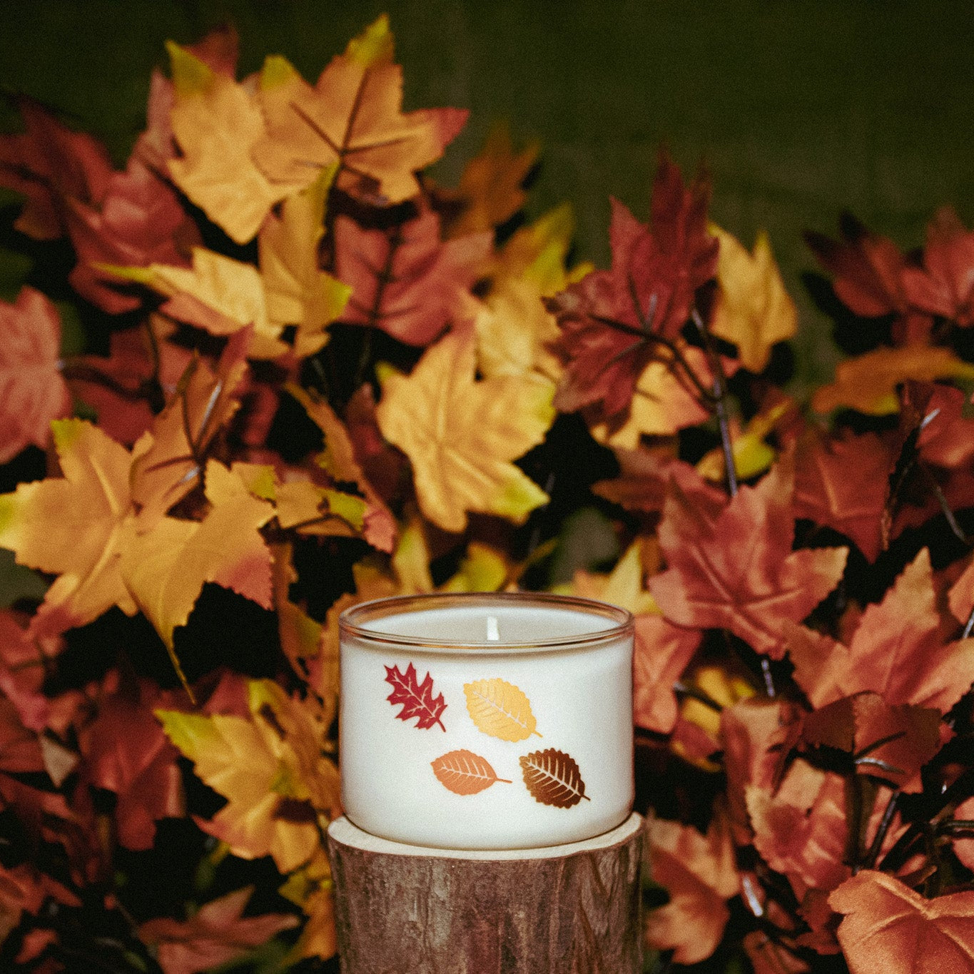 A white glass candle jar with a leaf pattern, displayed on a rustic wooden block surrounded by artificial autumn maple leaves.